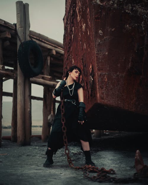 Woman in Black One Shoulder Dress and Fingerless Leather Gloves Dragging an Old Rusty Ship by its Anchor
