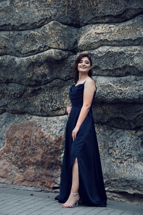 Free A young woman in a blue dress standing confidently by a rugged stone wall. Stock Photo