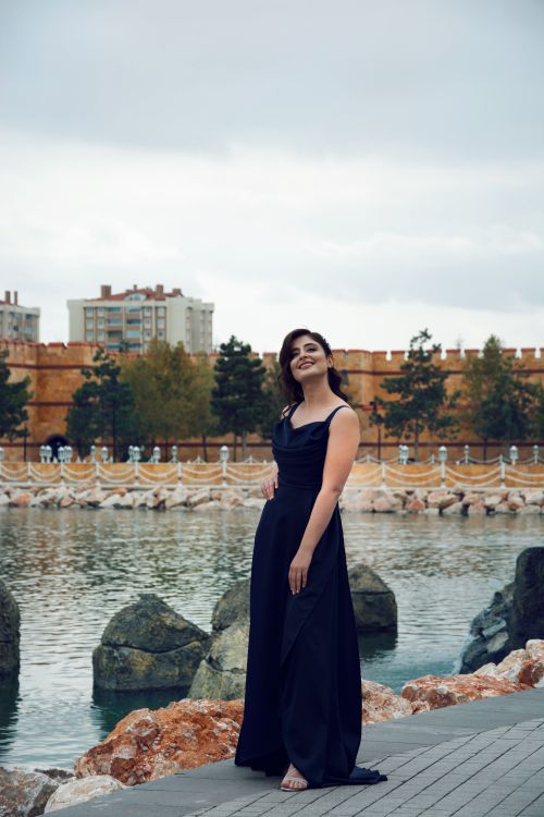 Free Stylish woman smiling in a black dress near an urban pond with a cityscape backdrop. Stock Photo