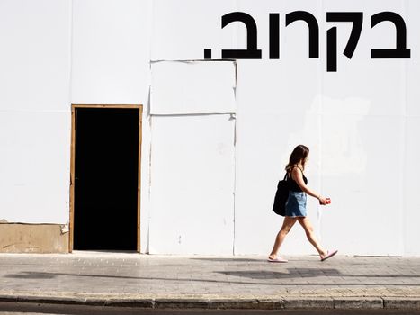 A woman walks by a white wall with Hebrew text in an urban street setting.