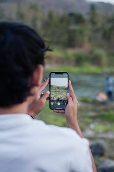 Back view of person using smartphone for outdoor photography by riverside.