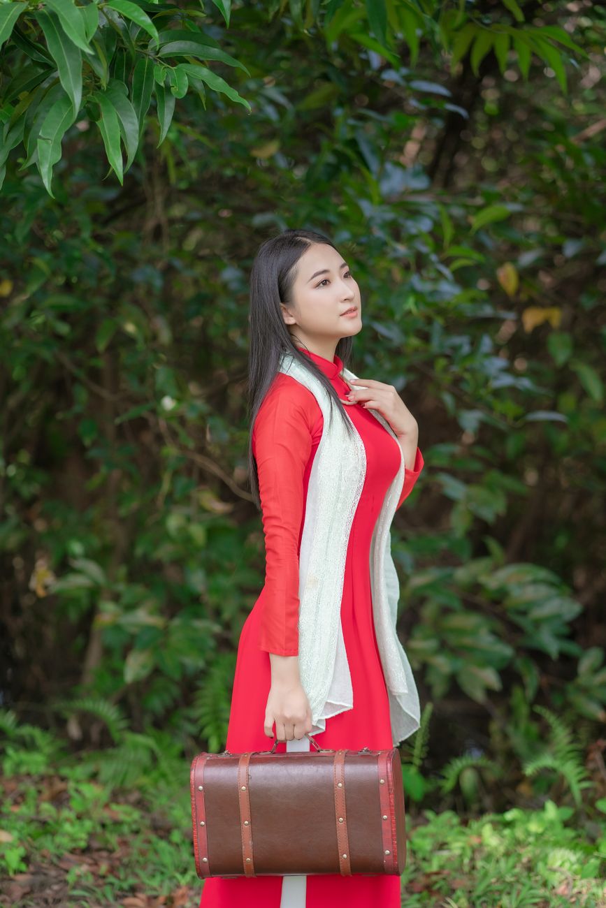 Elegant woman in traditional red ao dai with suitcase in a lush green setting.
