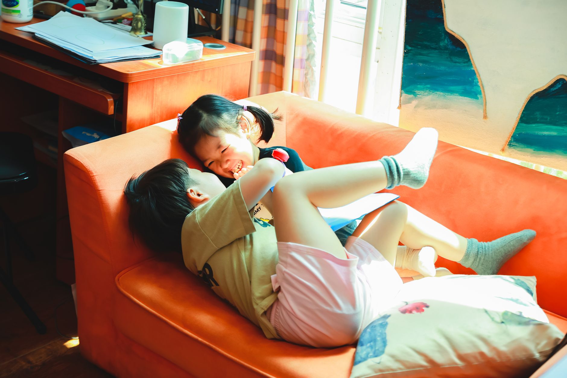 Two children joyfully playing and laughing on an orange sofa inside a cozy room.