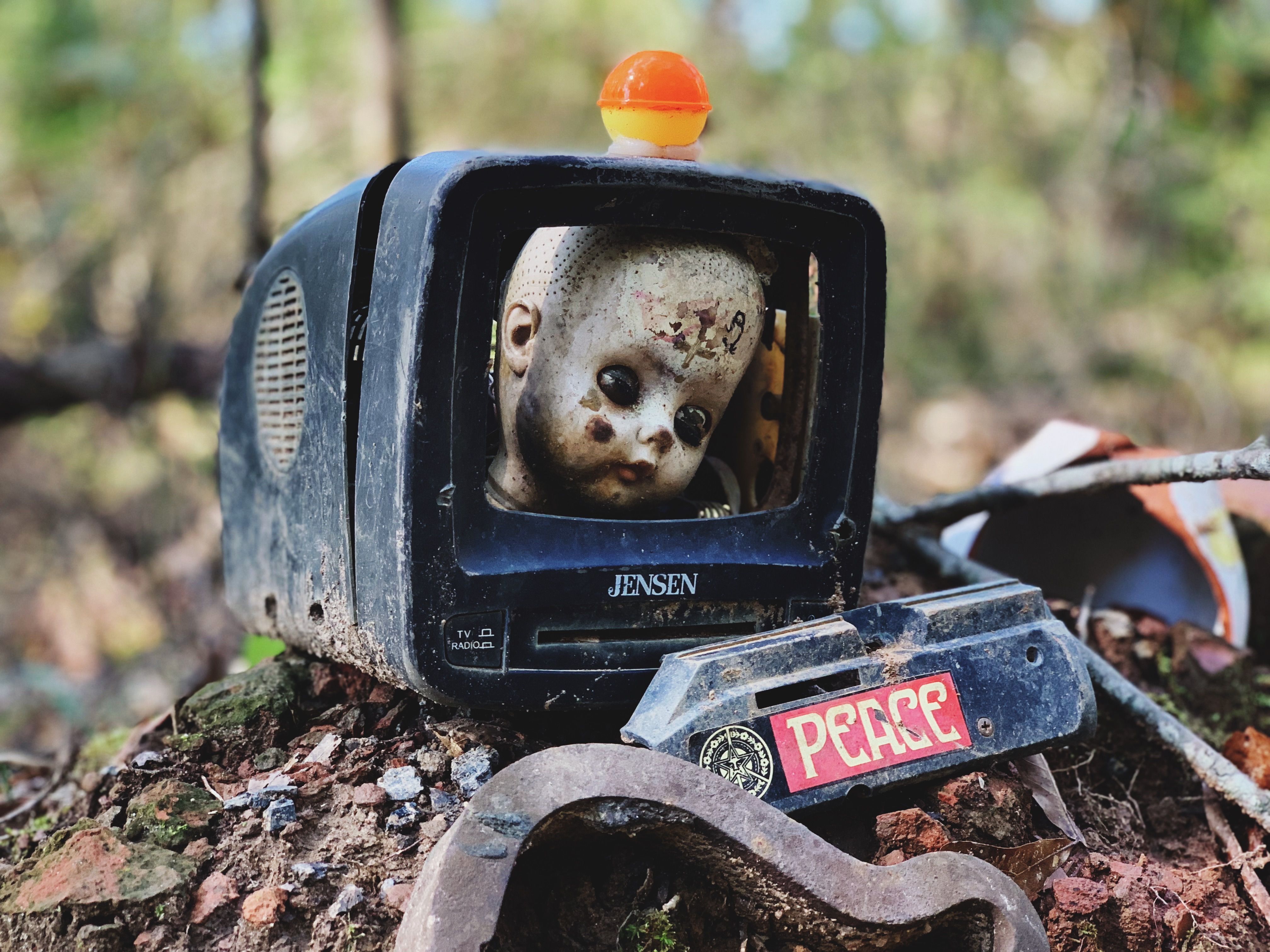 Rustic scene with a doll's head inside an old TV, featuring a peace sign, set outdoors.