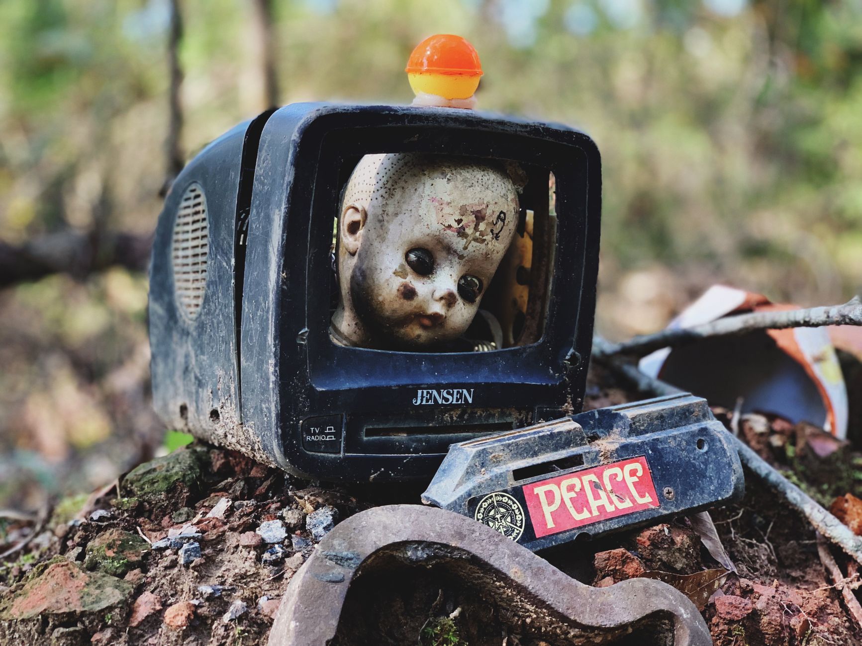Rustic scene with a doll's head inside an old TV, featuring a peace sign, set outdoors.