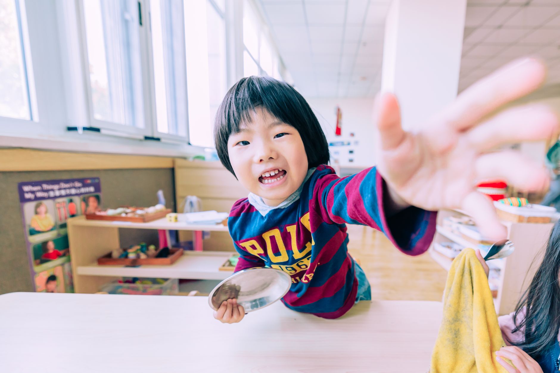 Joyful child reaching out towards the camera in a bright classroom, engaging and playful atmosphere.
