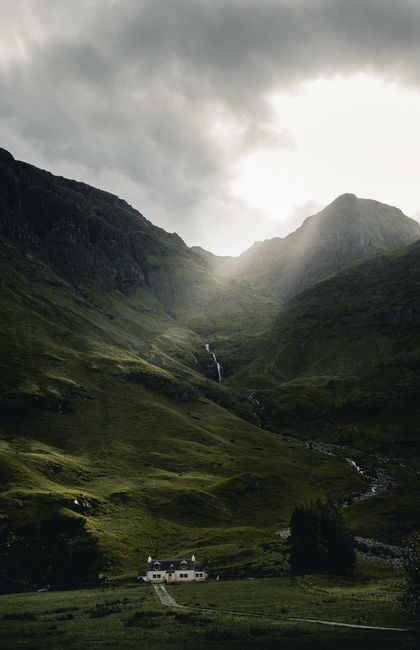 Breathtaking view of a cottage nestled in lush green Scottish highlands under dramatic, cloudy skies.