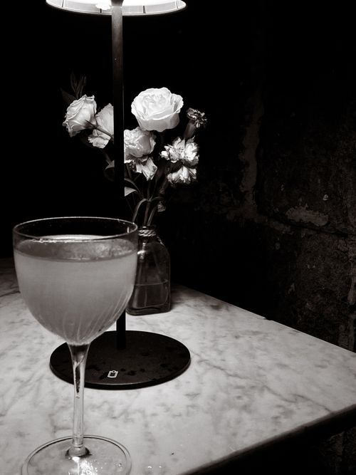 Free A stylish cocktail glass sits on a marble table beside a vase of flowers, captured in black and white. Stock Photo