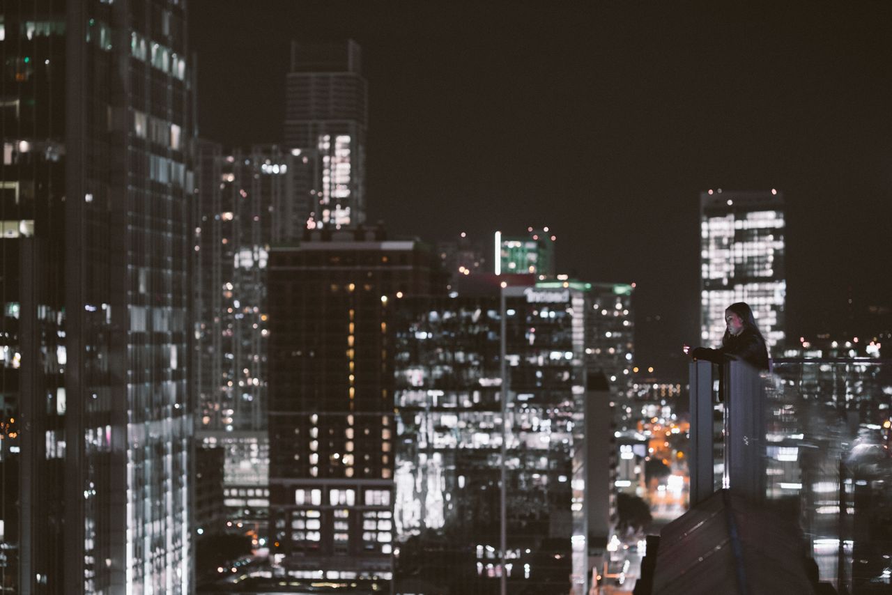 a person gazes over a bustling cityscape from a rooftop at night surro