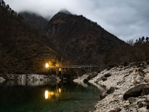 Free A moody landscape of a bridge and mountain under twilight, with lights reflecting on the calm lake. Stock Photo