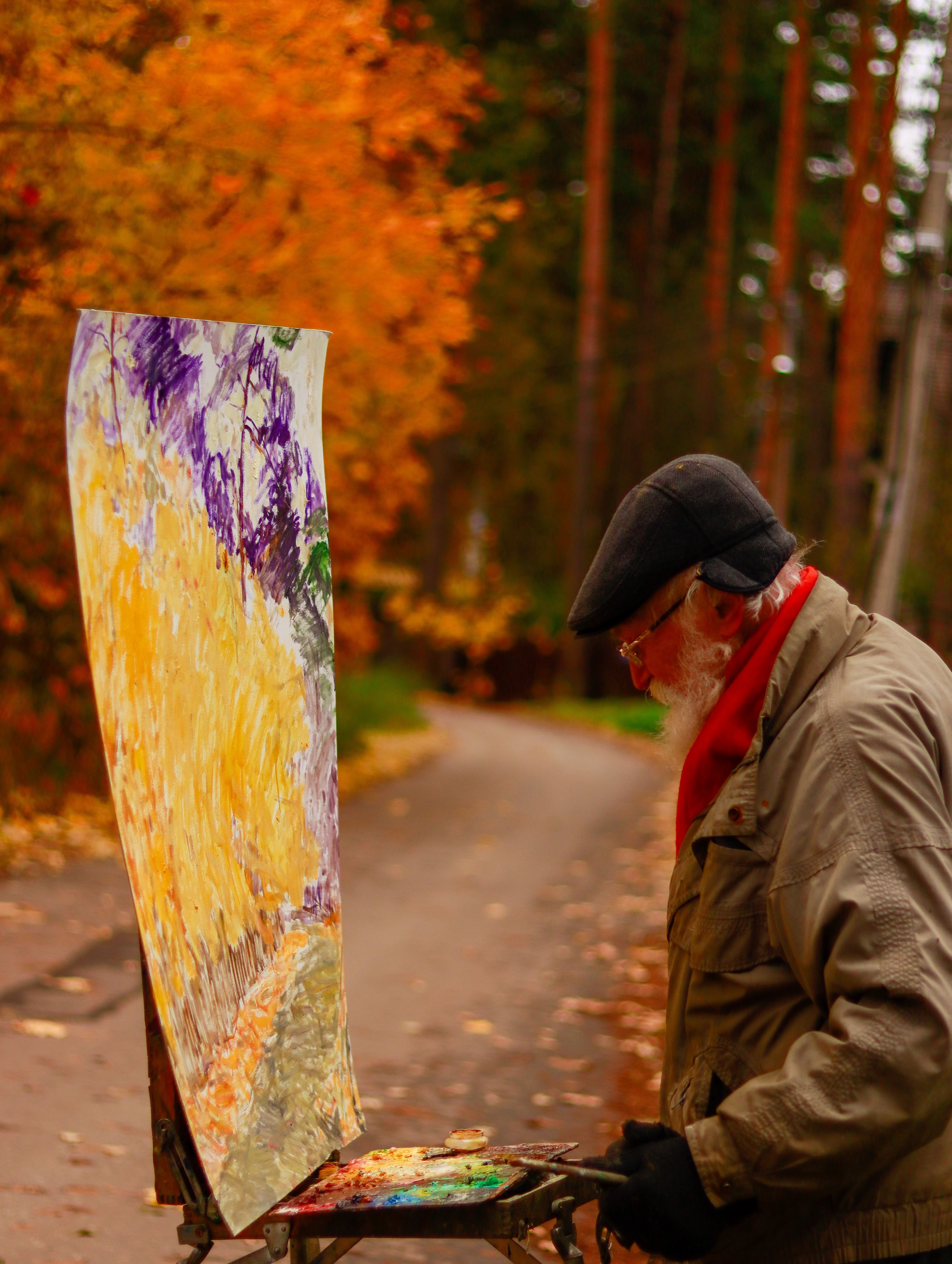 A senior artist paints an autumn scene in a park. Vibrant fall colors surround him.