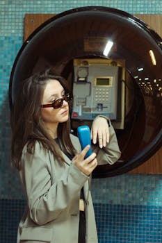 Stylish young woman in sunglasses using a vintage payphone against a tiled wall.