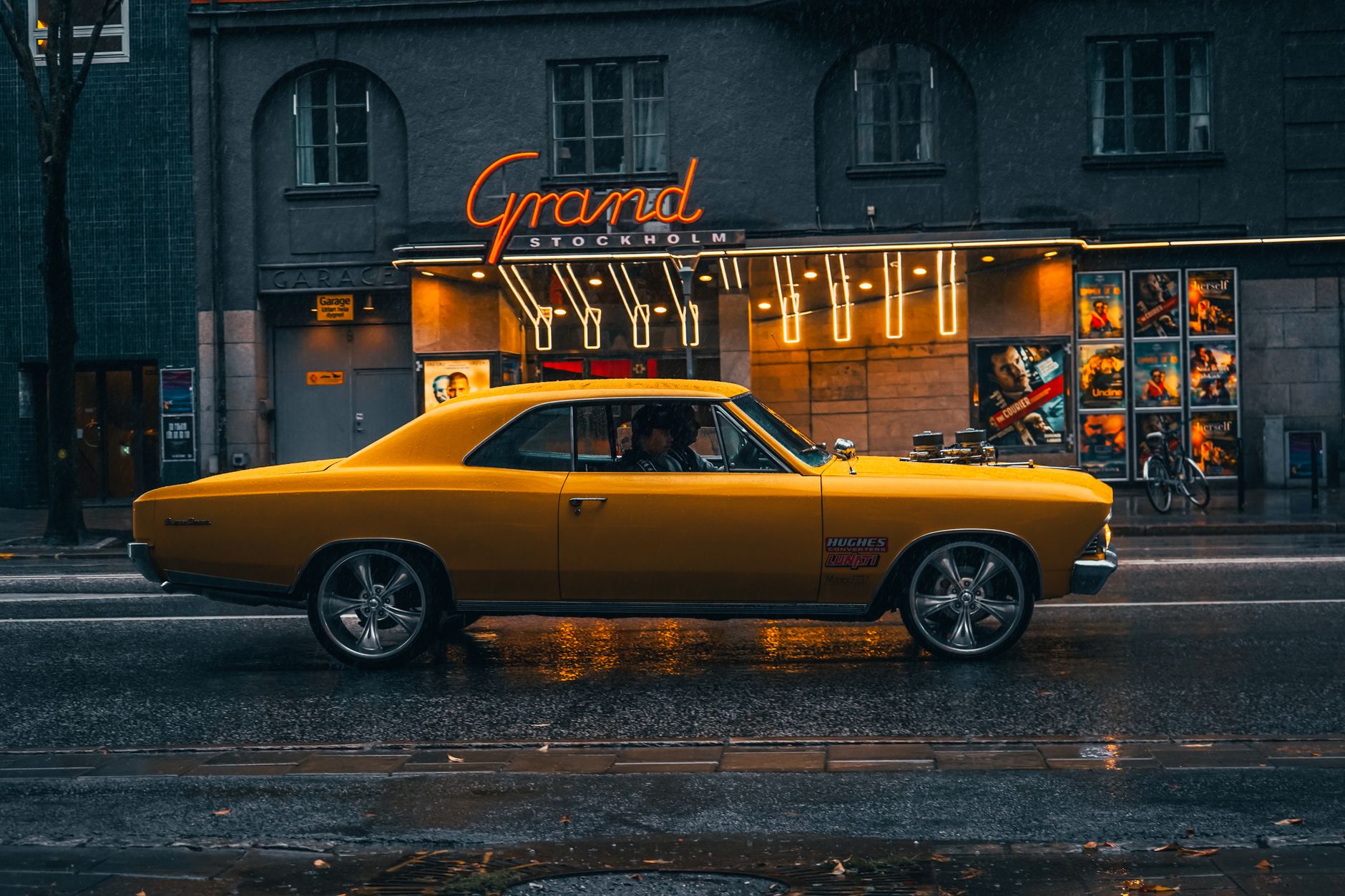 A vintage yellow muscle car cruises a rainy street outside a movie theater in Stockholm.