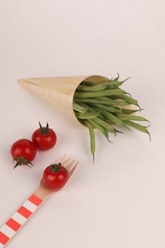 Minimalist still life featuring cherry tomatoes and green beans in a paper cone with a wooden fork.