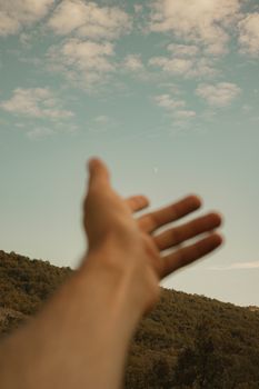 Blurred hand reaching towards a serene blue sky over a lush green mountain landscape.