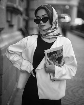 Stylish woman in sunglasses and headscarf holding magazines on a city sidewalk.