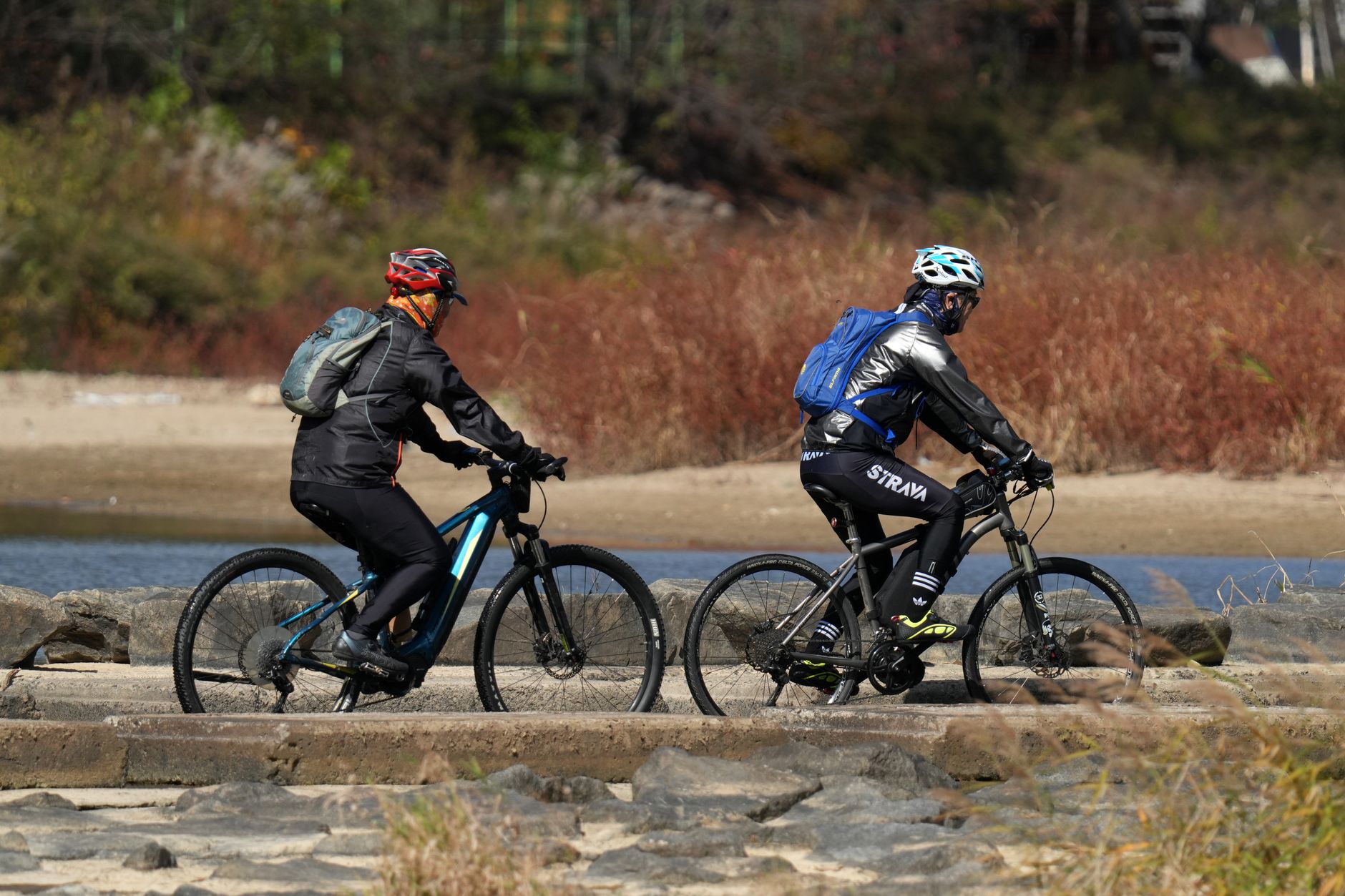Two cyclists ride along a scenic riverside trail with backpacks, enjoying outdoor recreation.