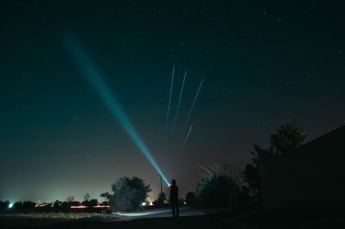 Free A man stands in a rural area, illuminating the night sky with a flashlight, under a starlit sky. Stock Photo