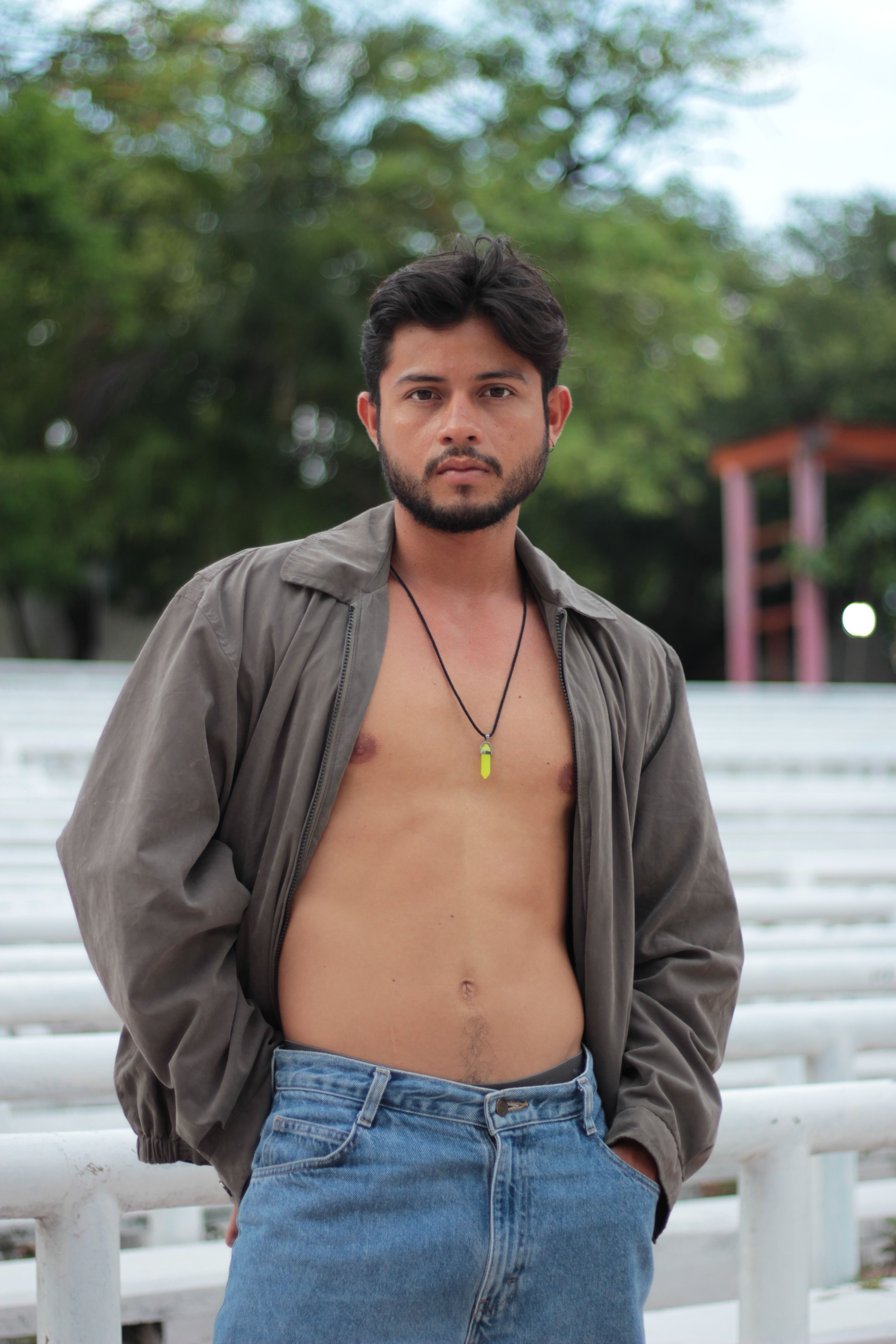 Shirtless young man with necklace posing outdoors in casual attire.