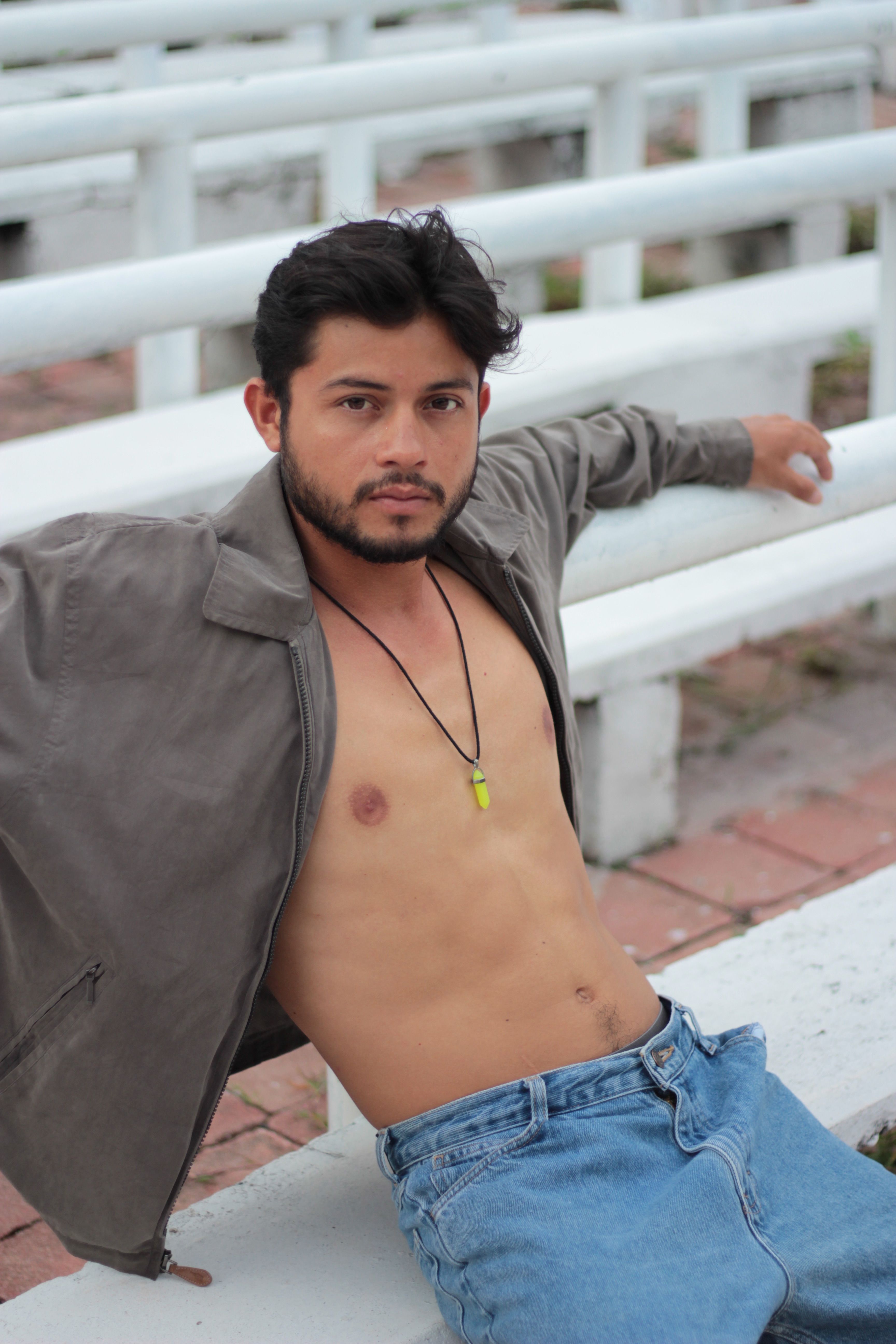 Shirtless man with beard and gray jacket sitting on white benches outdoors.