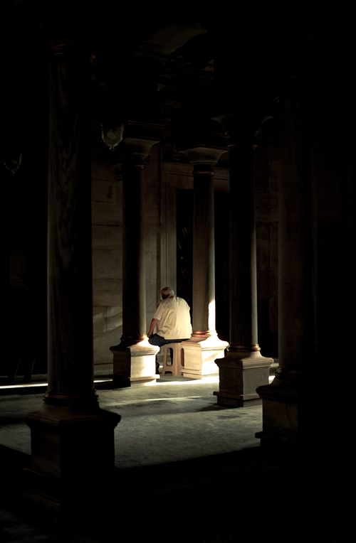 Free A solitary man sits in a dimly lit temple corridor, surrounded by tall columns. Stock Photo