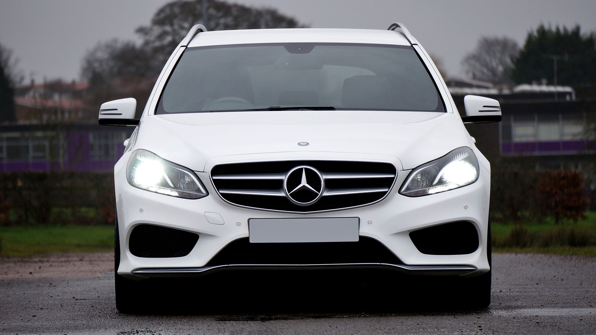 Luxury white Mercedes-Benz E-Class station wagon with headlights on, parked in an urban setting.