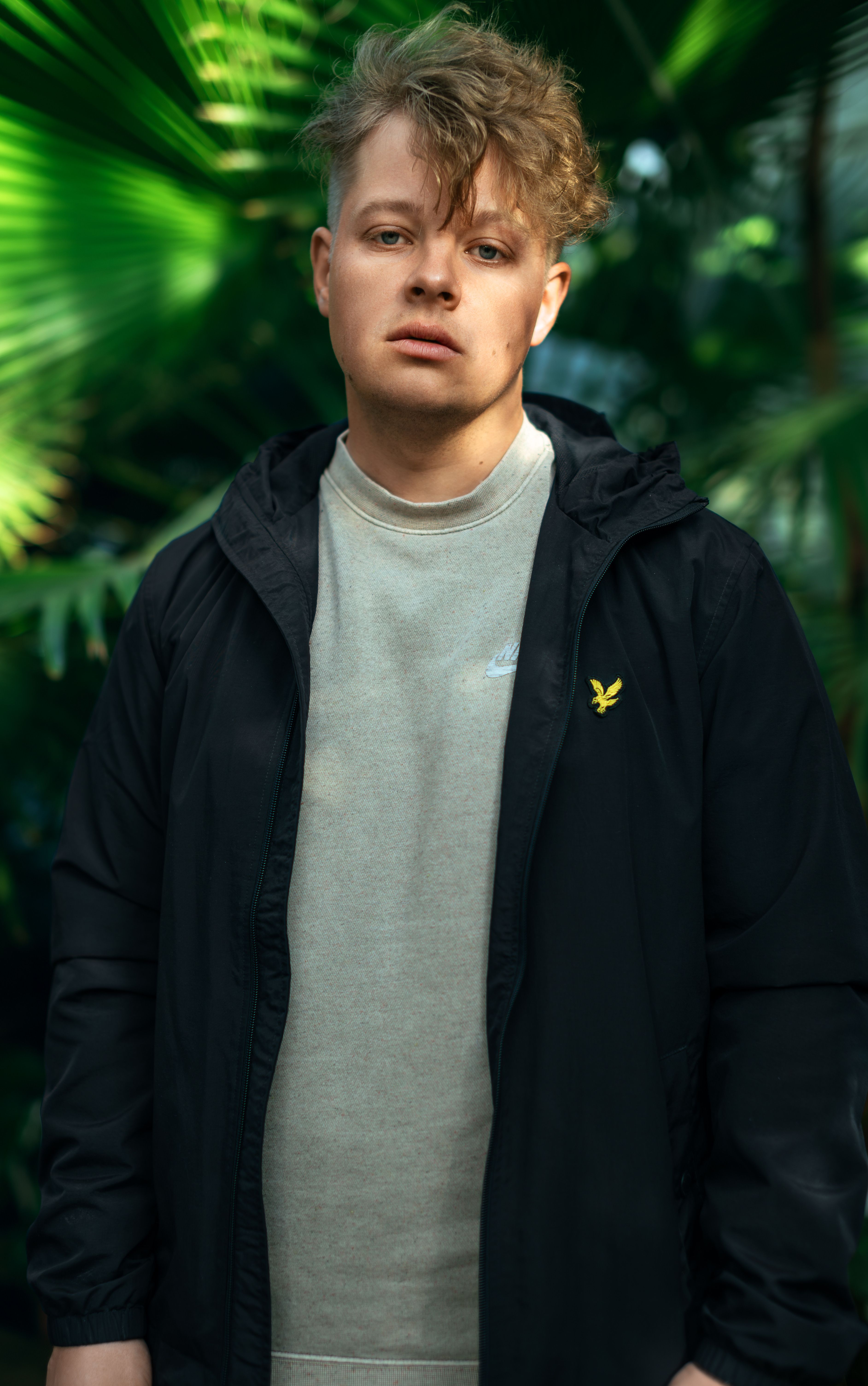 Portrait of a young man in a black jacket standing among lush green plants in a botanical garden.