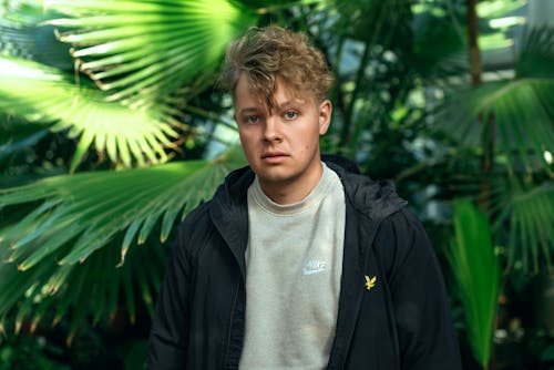 Young Man in a Jacket Standing on the Background of Plants in a Botanical Garden 