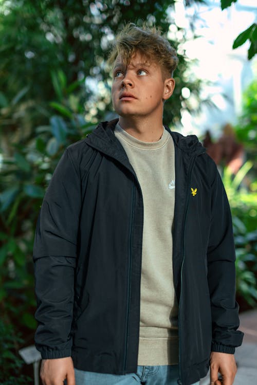 Young Man in a Sweatshirt and Jacket Standing on the Background of Plants 