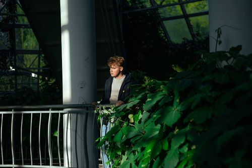 Free A young man stands on a balcony in a sunlit indoor garden surrounded by lush green plants. Stock Photo