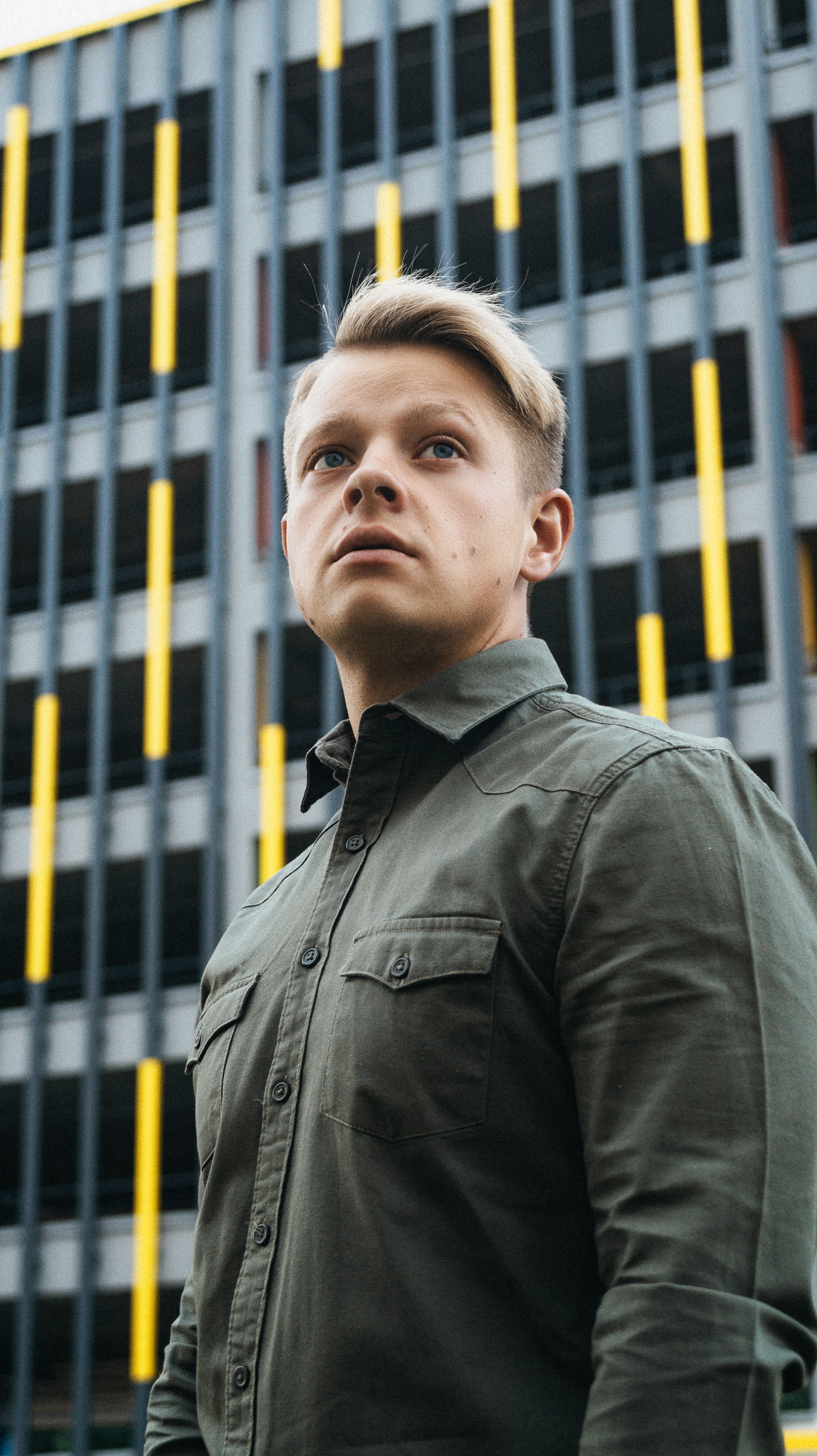 Young man in olive green shirt standing in front of urban architecture, looking off camera.