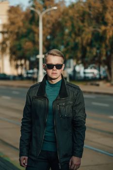 Fashionable man in a jacket and sunglasses standing confidently on a city street.