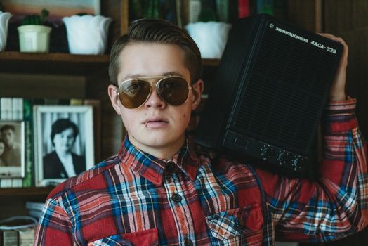 Teenager in plaid shirt and sunglasses holding a vintage speaker indoors.