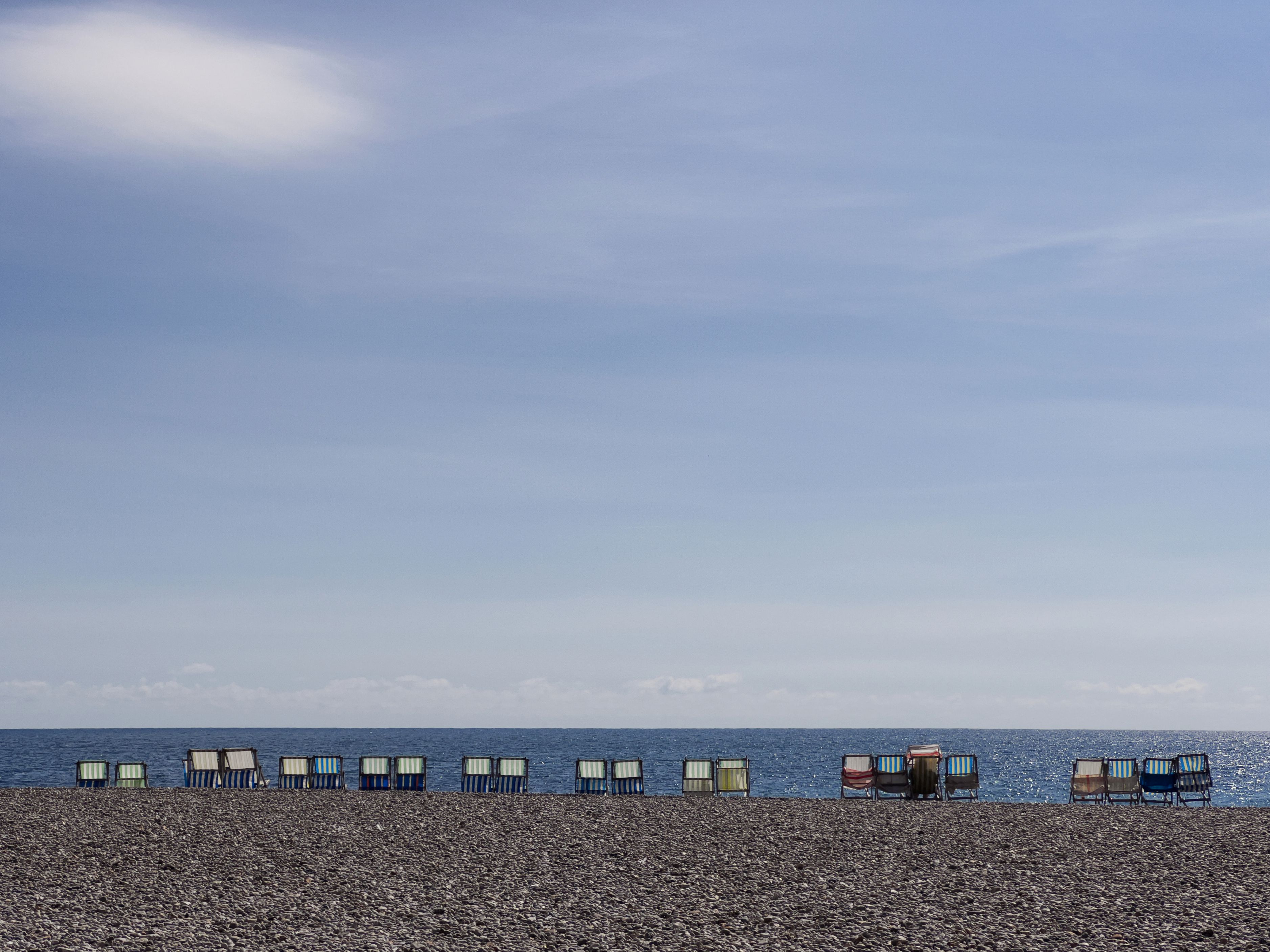 Empty deckchairs line a pebble beach under a vast blue sky by the sea.