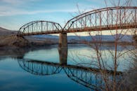 一座质朴的钢桥下，光秃秃的树木倒映在晴朗天空下的宁静河流中