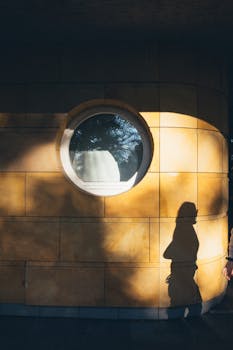 Sunlit yellow wall with round window and shadow of a woman walking.