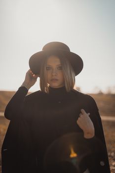 Elegant portrait of a woman in black clothing and hat at sunset, highlighting fashion and style.