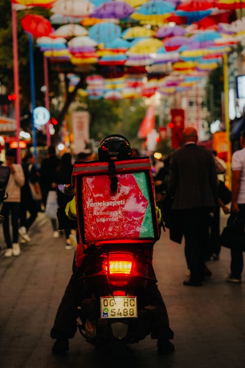 Back of a Delivery Person Riding a Motor Scooter across a Busy Street