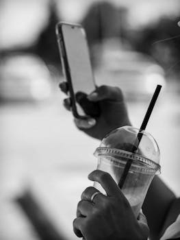 Black and white photo of hands holding smartphone and plastic cup with straw.