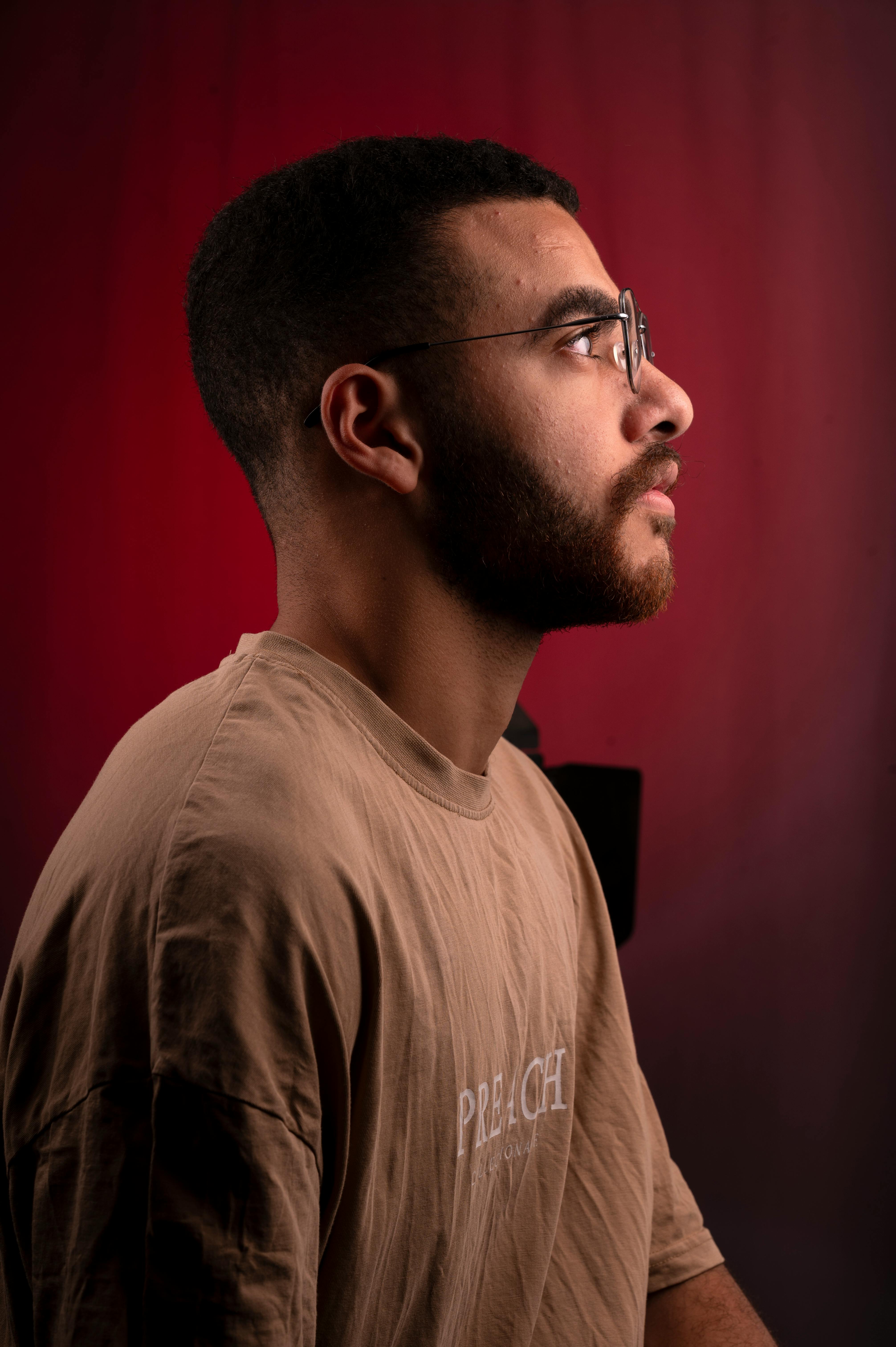 Dramatic side profile of a young man with a beard wearing glasses and a t-shirt on a red background.