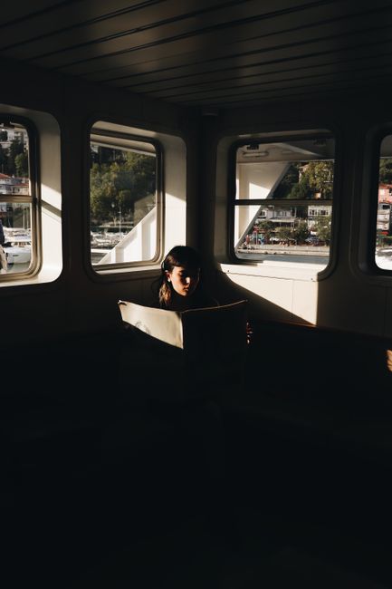A woman reads a newspaper on a ferry, bathed in soft sunlight with a scenic view outside.