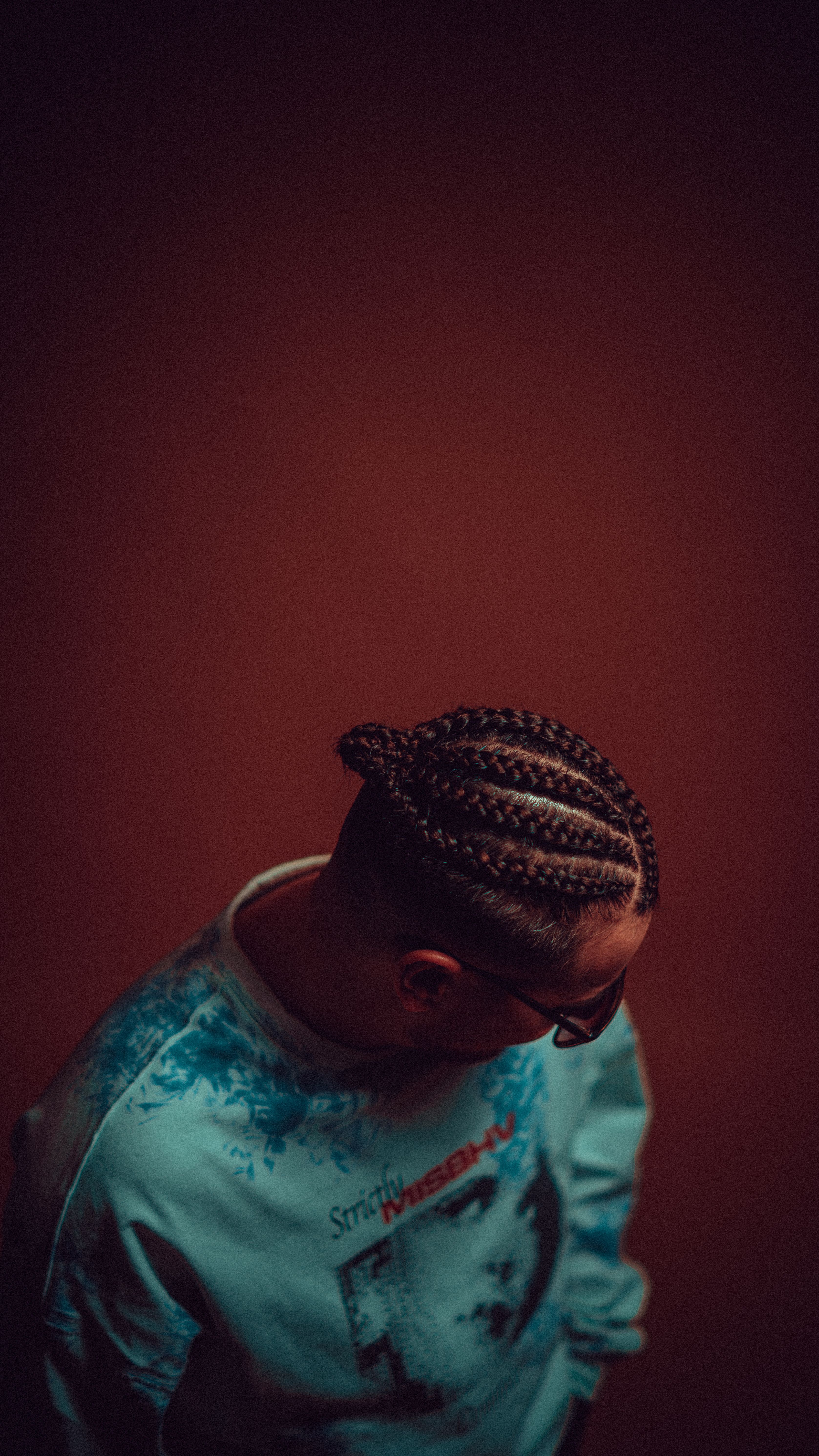 Studio portrait of a man with braided hair in a light blue sweatshirt. Dark tones, obscured face.