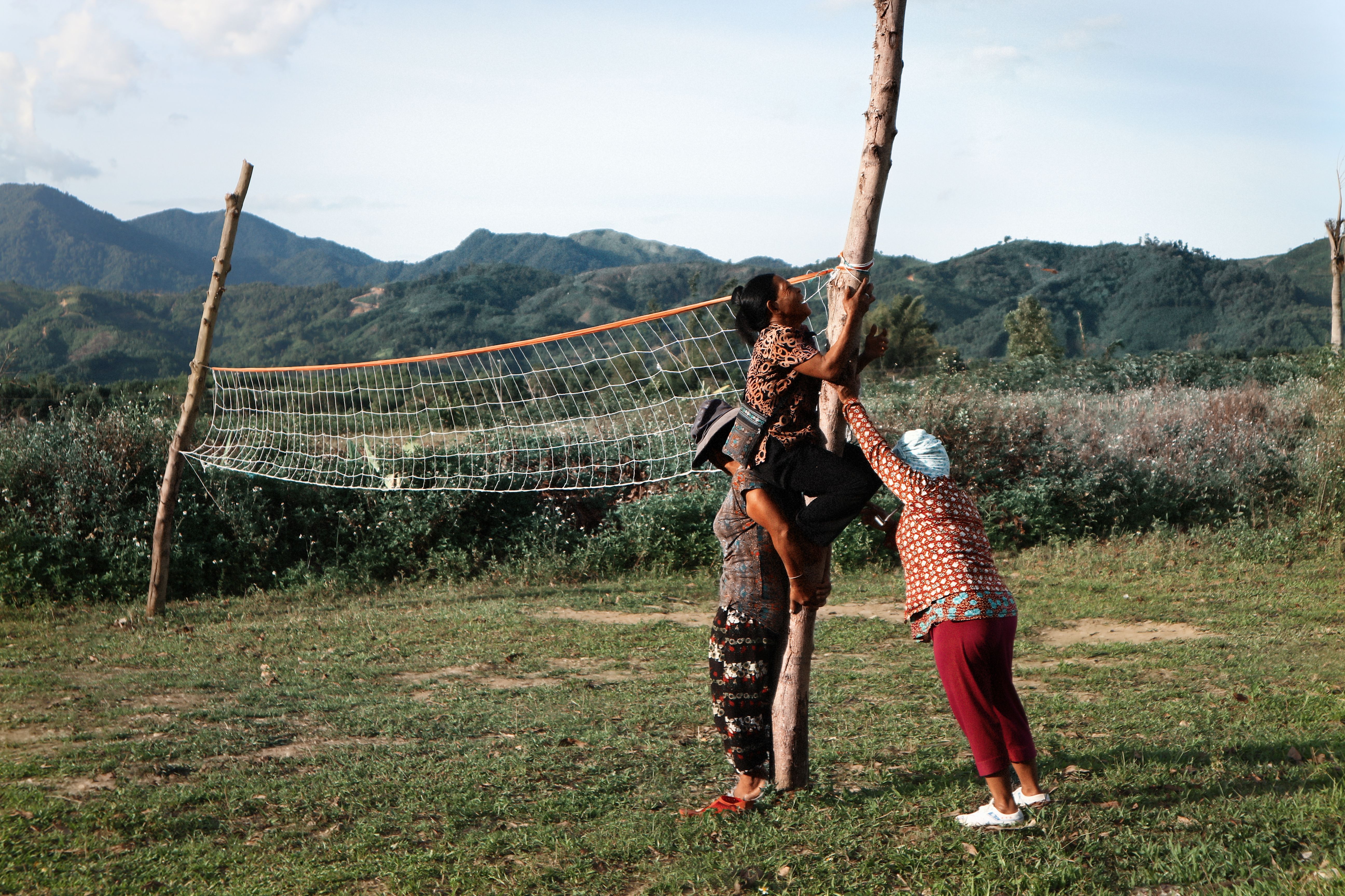 Women collaborating in a rural field to set up a volleyball net against mountain scenery.