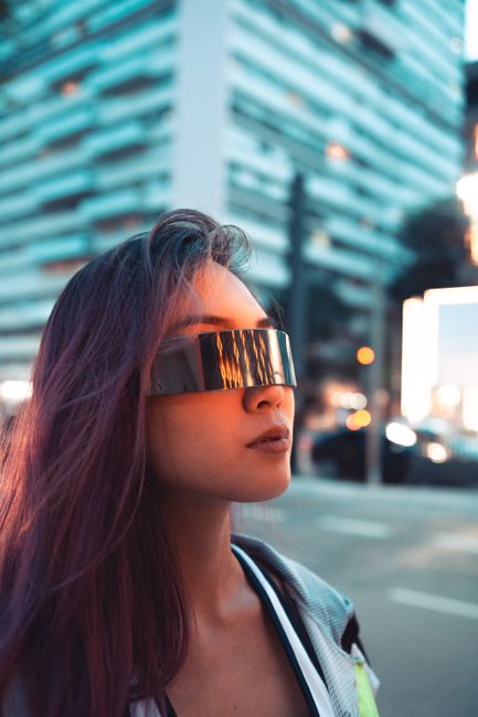 A young woman with futuristic sunglasses poses in an urban setting, exuding modern style.