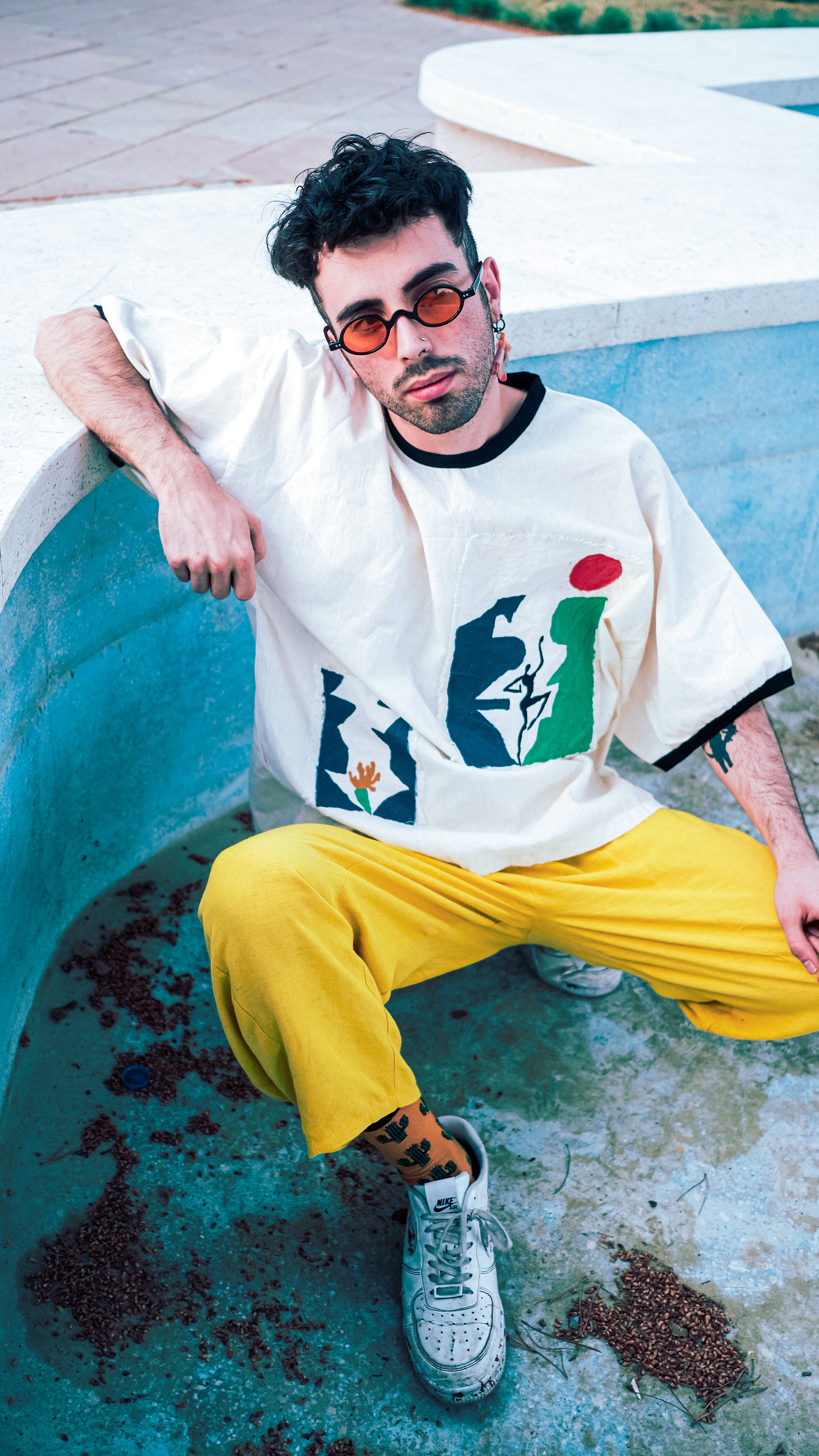Stylish young man posing fashionably in an empty pool wearing colorful attire and sunglasses.