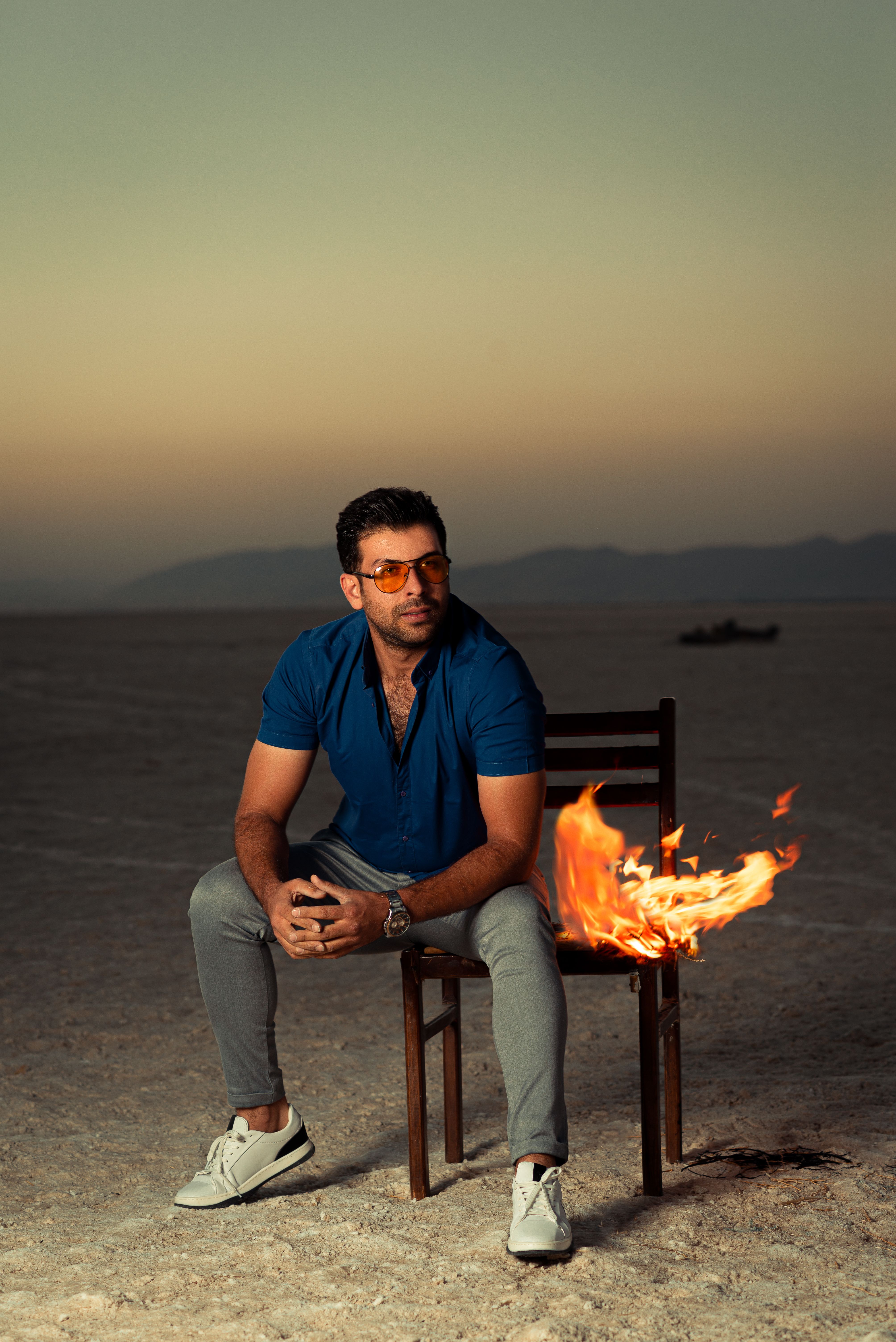 A man sits on a chair with flames in a desert during sunset, creating a dramatic scene.