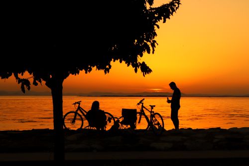 Free Silhouettes of cyclists relaxing by a lake during a vibrant sunset, creating a serene and picturesque scene. Stock Photo