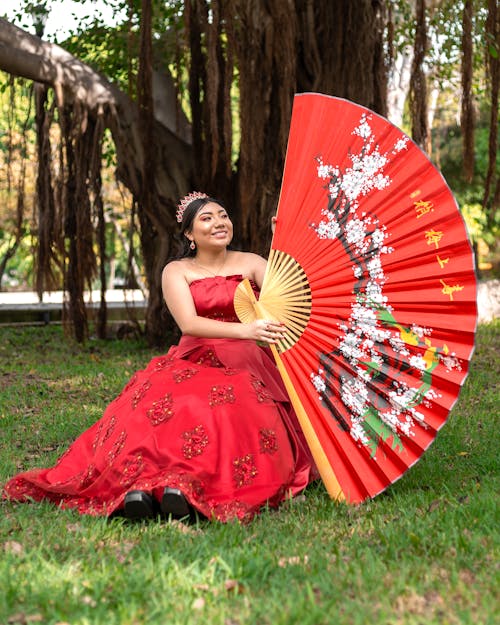 Young Woman in a Red Dress Holding a Large Red Fabric Fan Sitting in a Park 