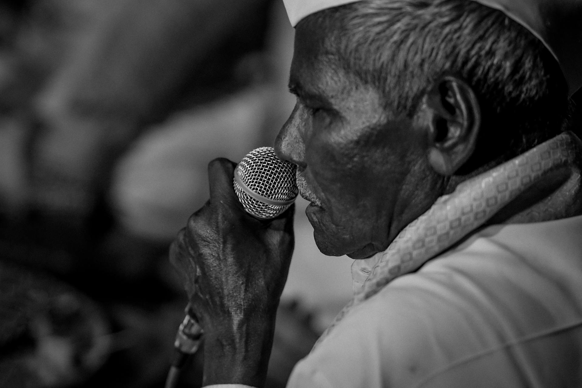 Black and white portrait of a senior Indian man in Surat speaking with a microphone.
