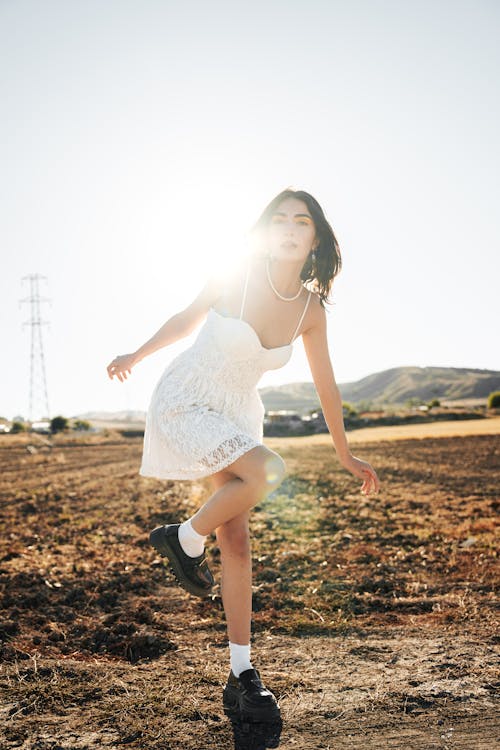 Young Model in a White Lace Mini Dress Standing on One Leg in a Field with the Sun Behind Her